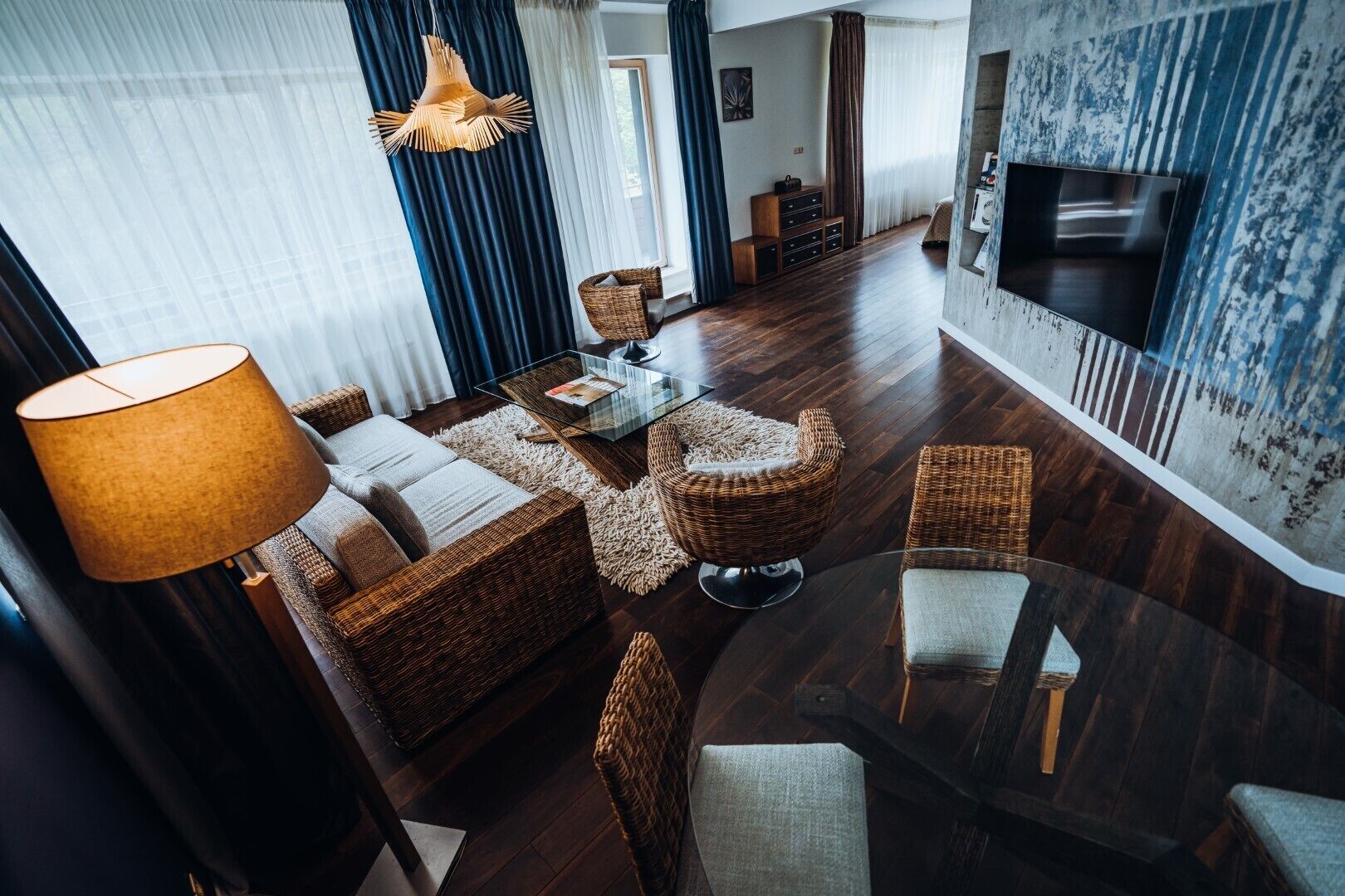 A modern living room with a sofa set, a glass coffee table, and wicker chairs. There is a TV mounted on the wall and a lamp on a side table.