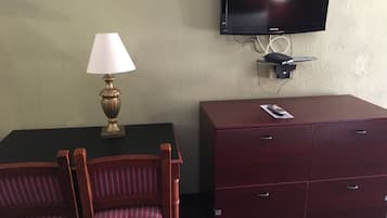 A hotel room with a TV mounted on the wall, a desk with a lamp, a dresser, and two chairs.