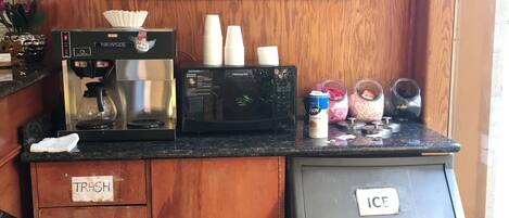 A kitchen area with a coffee machine, a microwave, and a refrigerator. There is a trash bin labeled "TRASH" and an ice bin labeled "ICE". A framed painting of a canal scene hangs above the counter.
