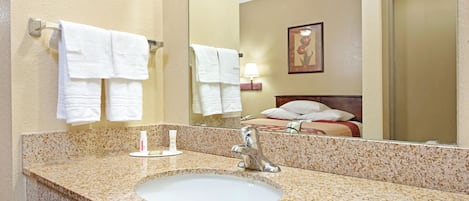 A hotel bathroom with a granite countertop sink, two white towels hanging on a towel rack, a mirror reflecting a bed with a pillow, and a framed picture on the wall.
