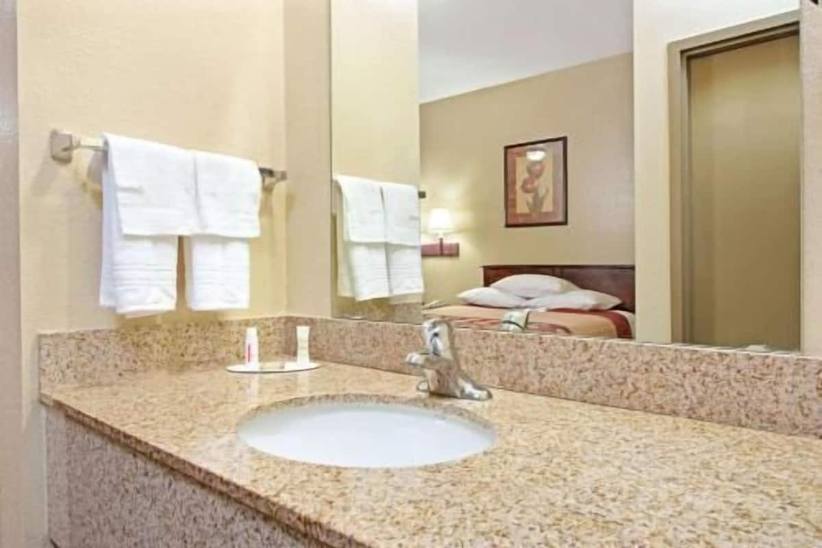Un baño de hotel con lavamanos de encimera de granito, un espejo y una cama con cabecero al fondo.