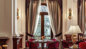 A dining area with a round table, red upholstered chairs, a chandelier, and a window with curtains. - Grecotel Larissa Imperial (Larissa)