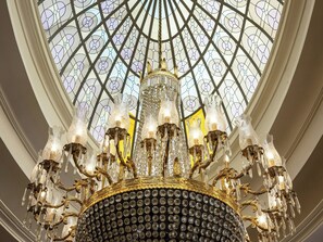 Un magnifique lustre orné de décorations en cristal et de motifs dorés, qui se détache sur un vaste plafond constitué de vitraux complexes.