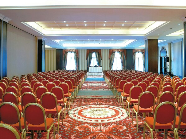 Un salón de conferencias con filas de sillas, piso alfombrado y un escenario al frente.