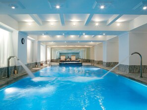 Une grande piscine intérieure dotée d’un espace de repos central et de plusieurs luminaires au plafond.
