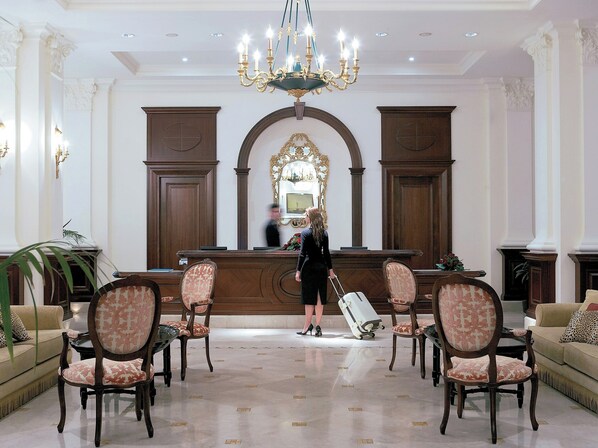 Un hall con mostrador de recepción, sillas, una araña de cristal y un espejo grande.