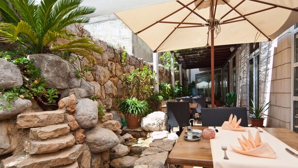 A stone wall with plants, a wooden table with a tablecloth, and an umbrella. - Hotel Perla (Dubrovnik)