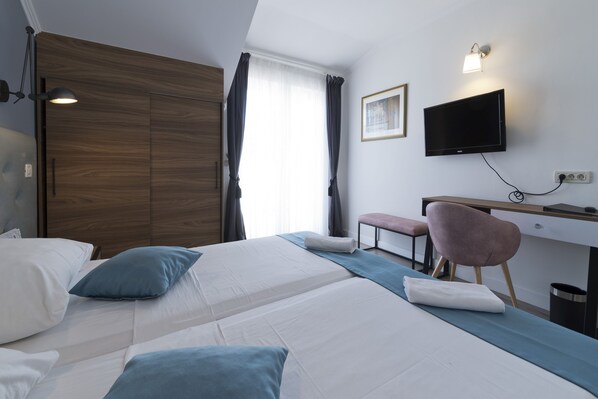 A hotel room with two beds, a desk, a chair, a TV, and a window with curtains. - Hotel Perla (Dubrovnik)