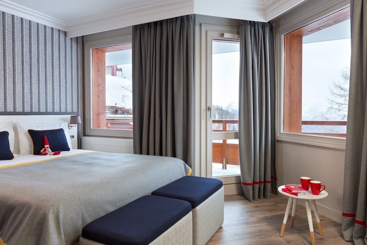 Une chambre d’hôtel avec un grand lit, une petite table avec des tasses rouges et une vue sur un paysage enneigé par la fenêtre.