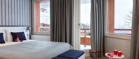 Une chambre d’hôtel avec un grand lit, une petite table avec des tasses rouges et une vue sur un paysage enneigé par la fenêtre.