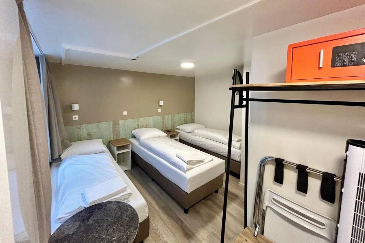 Ein Hotelzimmer mit zwei Einzelbetten, einem Etagenbett, einem kleinen Tisch, einem Heizkörper und einem Fenster mit Vorhängen.