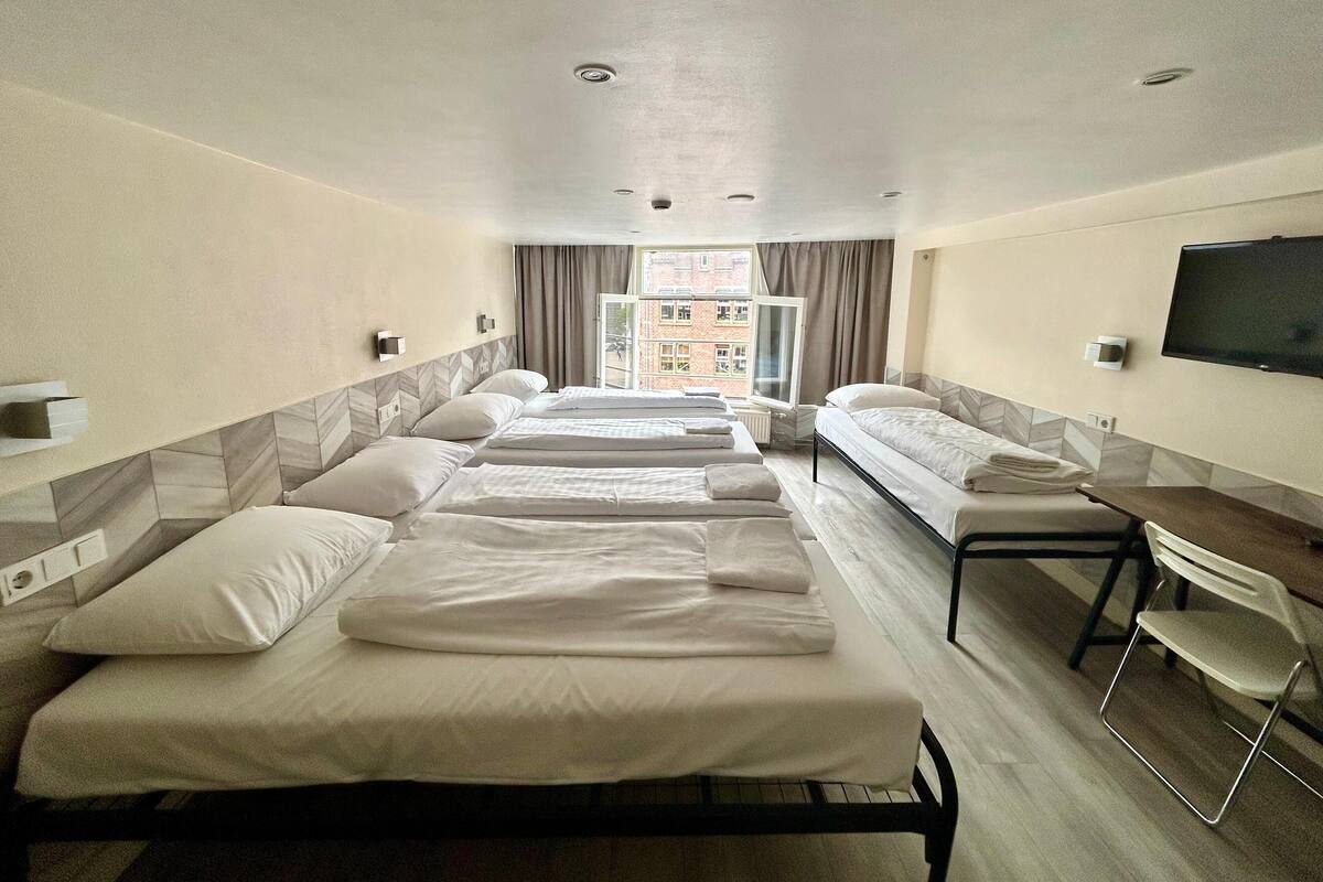 Ein Hotelzimmer mit drei Einzelbetten, einem Schreibtisch, einem Stuhl, einem Fernseher und einem Fenster mit Vorhängen.