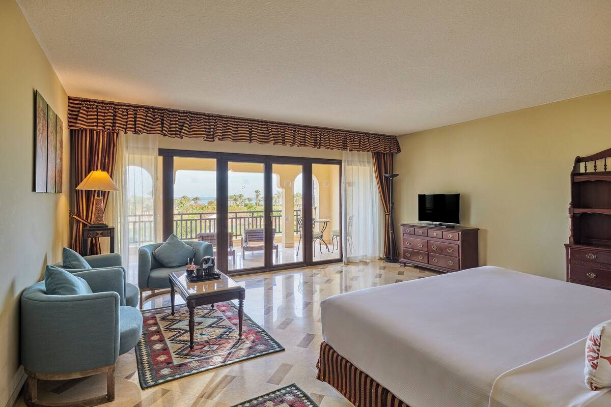 Une chambre d’hôtel dotée d’un grand lit, d’un coin salon avec deux fauteuils et une table basse, d’une télévision et d’un balcon offrant une vue agréable.