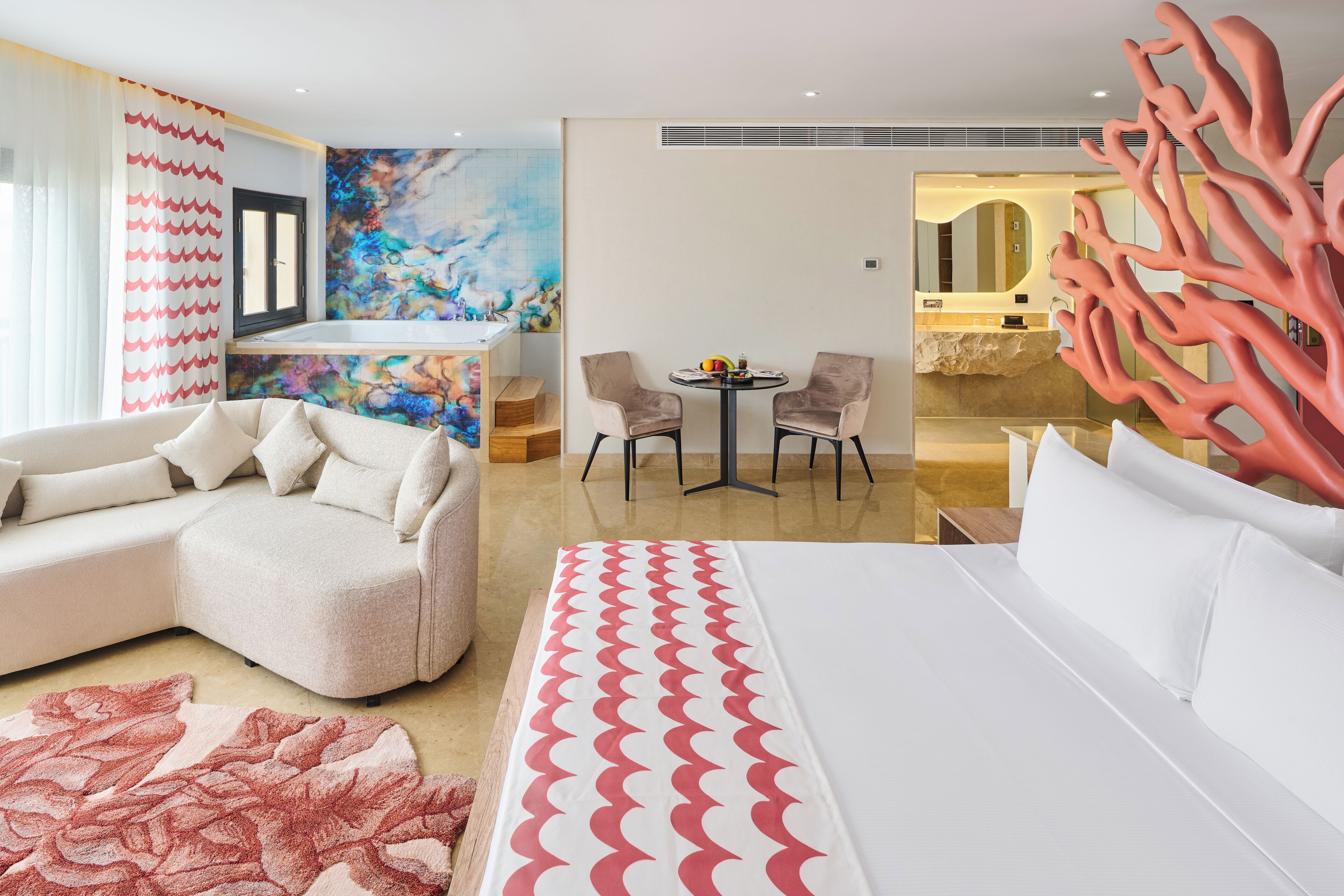 A modern hotel room with a bed, a sofa, a dining table with chairs, a bathroom with a mirror, and a colorful abstract painting on the wall.