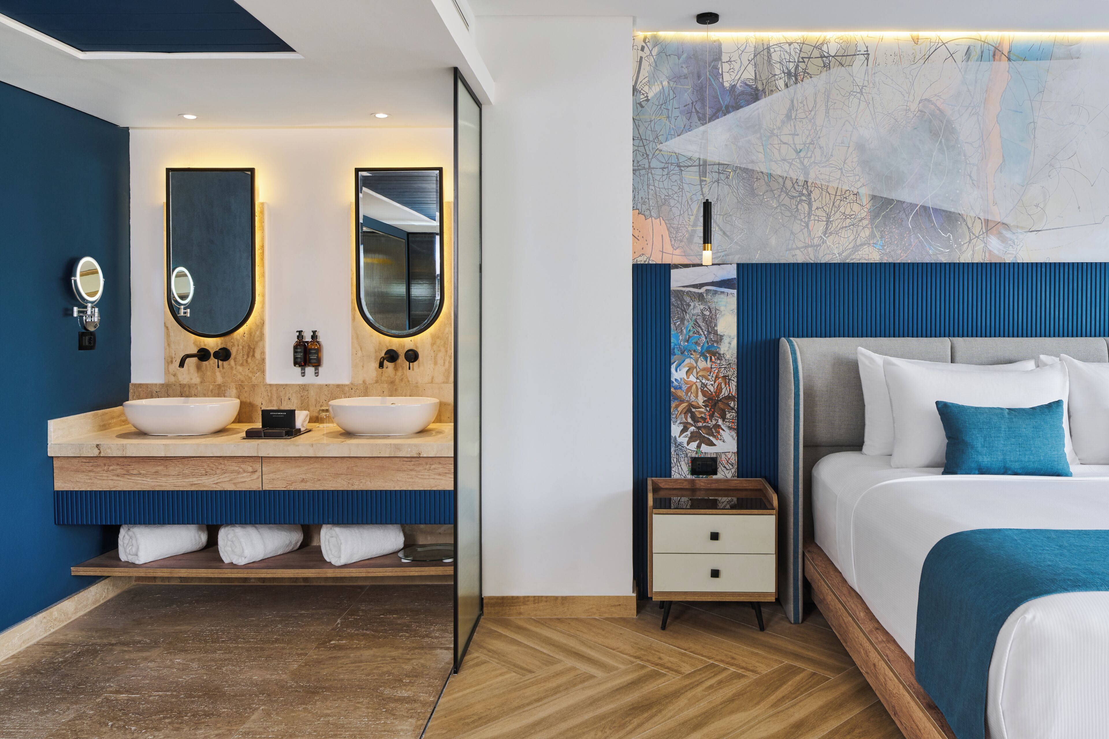 A modern hotel room with a wooden vanity, two sinks, a large mirror, a bed with a blue accent, and a wall-mounted artwork.
