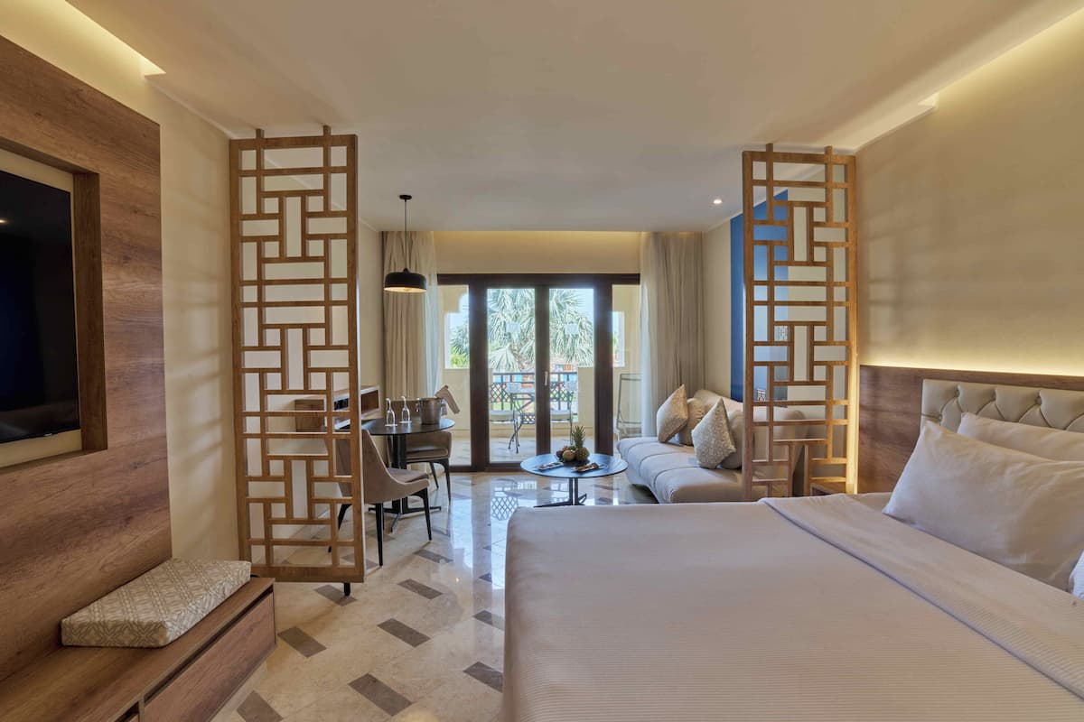 Une chambre d’hôtel dotée d’un grand lit, d’un meuble en bois pour la télévision, d’un bureau avec des chaises, d’un canapé et d’un balcon offrant une vue sur les palmiers.