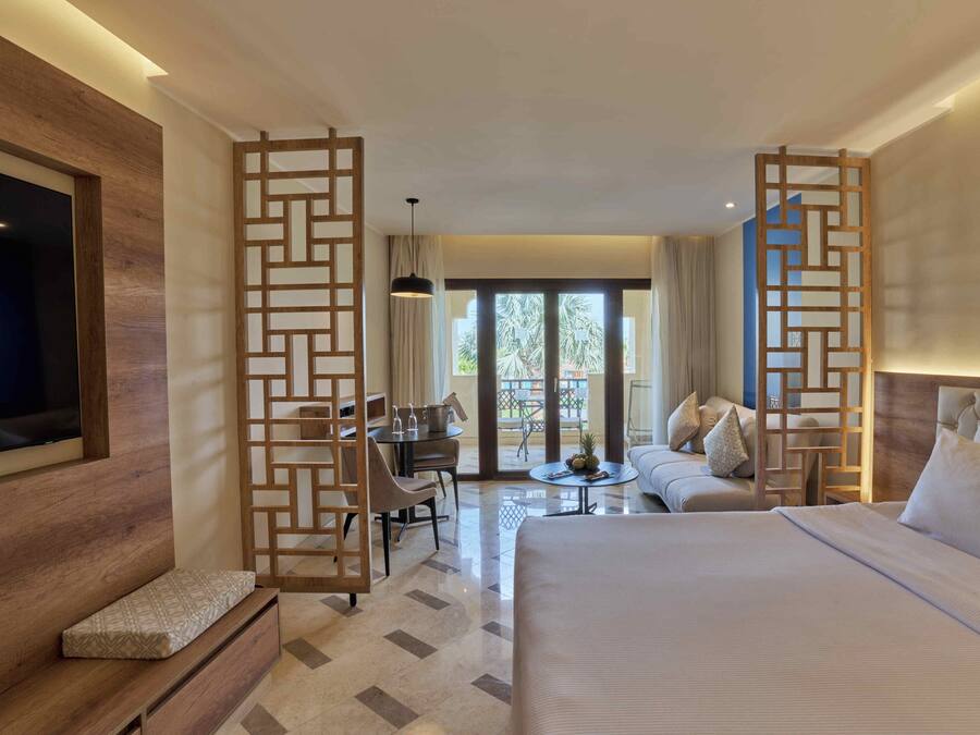 A hotel room with a large bed, a wooden TV stand, a desk with chairs, a sofa, and a balcony with a view of palm trees.