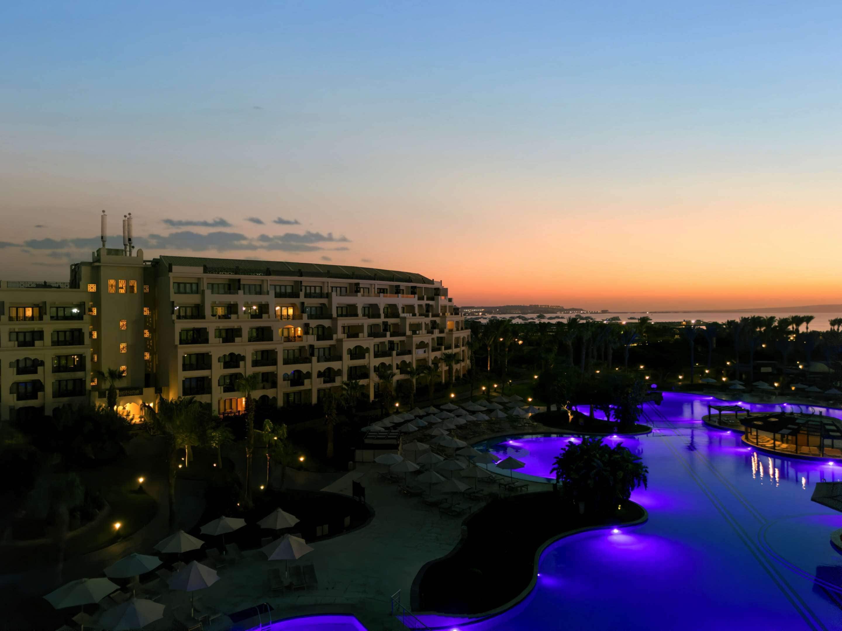 A hotel with multiple floors, a swimming pool with blue lighting, and a view of the sunset over the ocean.
