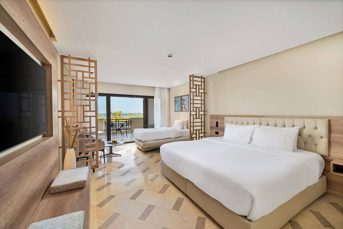 Une chambre d’hôtel avec un grand lit, deux petits lits, une télévision, un balcon avec une table et des chaises, et un paravent décoratif en bois.