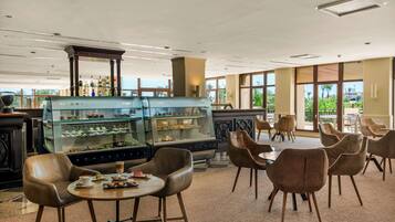 A spacious cafe with leather chairs, a dessert display case, and large windows offering a view of greenery.