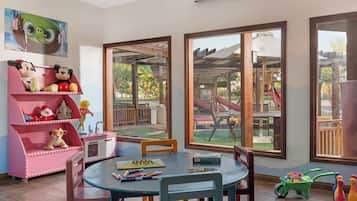 Une salle de jeux pour enfants avec une grande fenêtre, une maison de poupée rose et une table à manger avec des chaises.