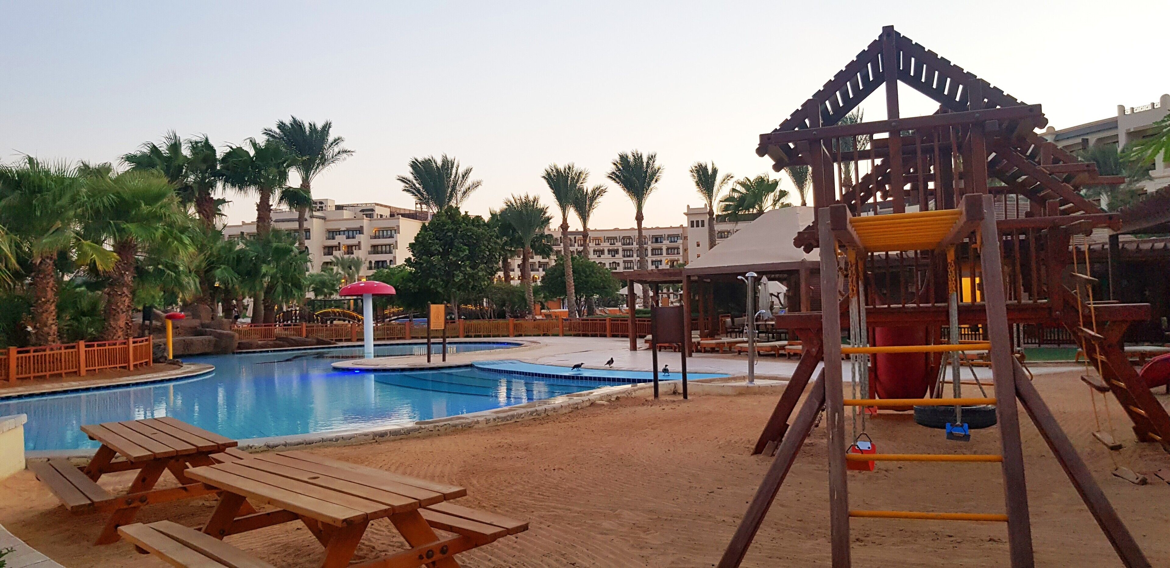 A pool area with wooden picnic tables, a playground structure, and palm trees.