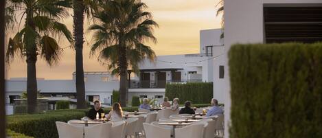 An outdoor dining area with tables and chairs, palm trees, and a building in the background.