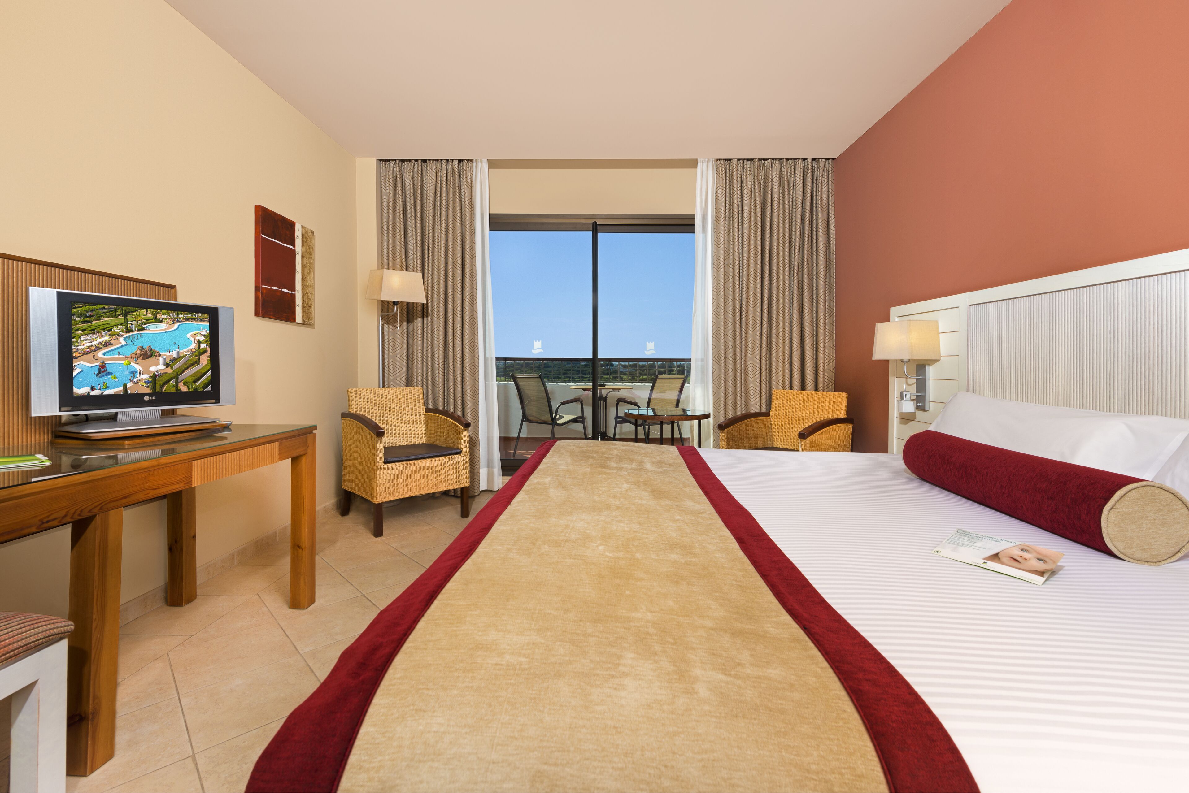 Een hotelkamer met een bed, een bureau met televisie, twee stoelen, een raam met uitzicht en een rood tapijt.
