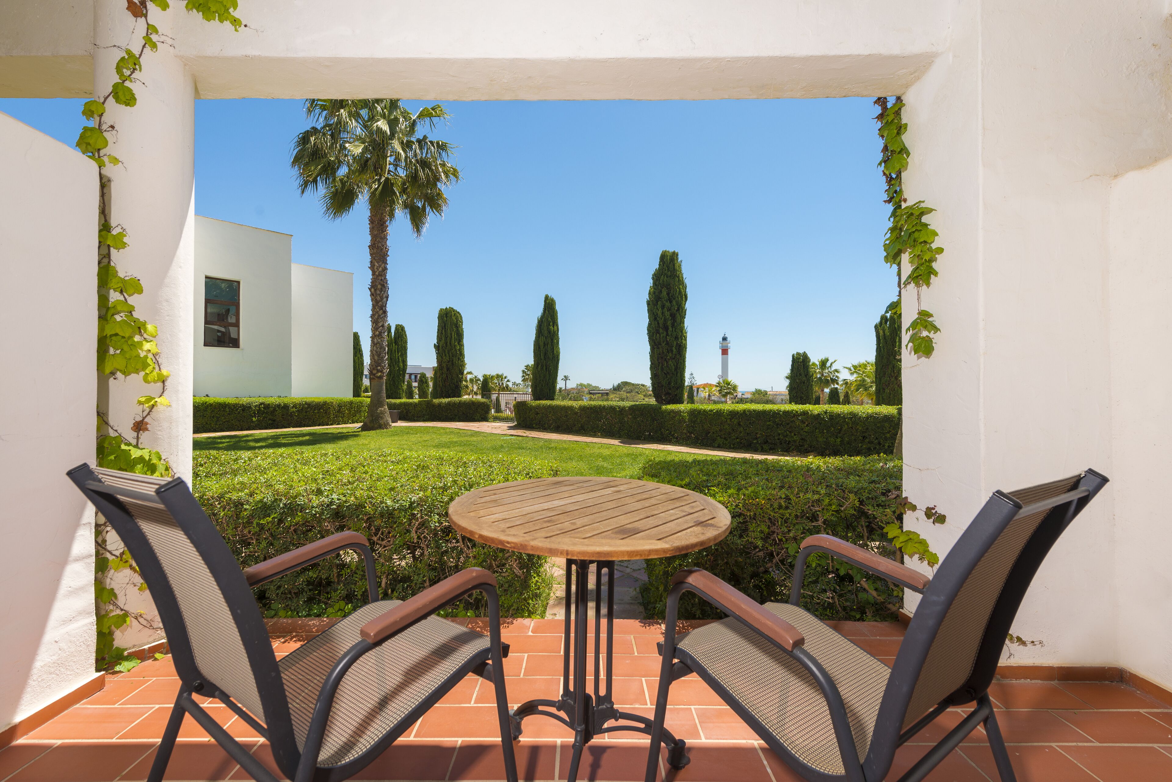 Un patio con dos sillas y una mesa redonda, con vistas a un jardín cuidado, con una palmera y setos.