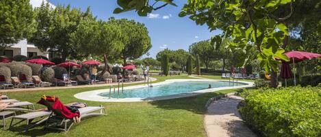 A pool area with lounge chairs and umbrellas, surrounded by greenery and trees.