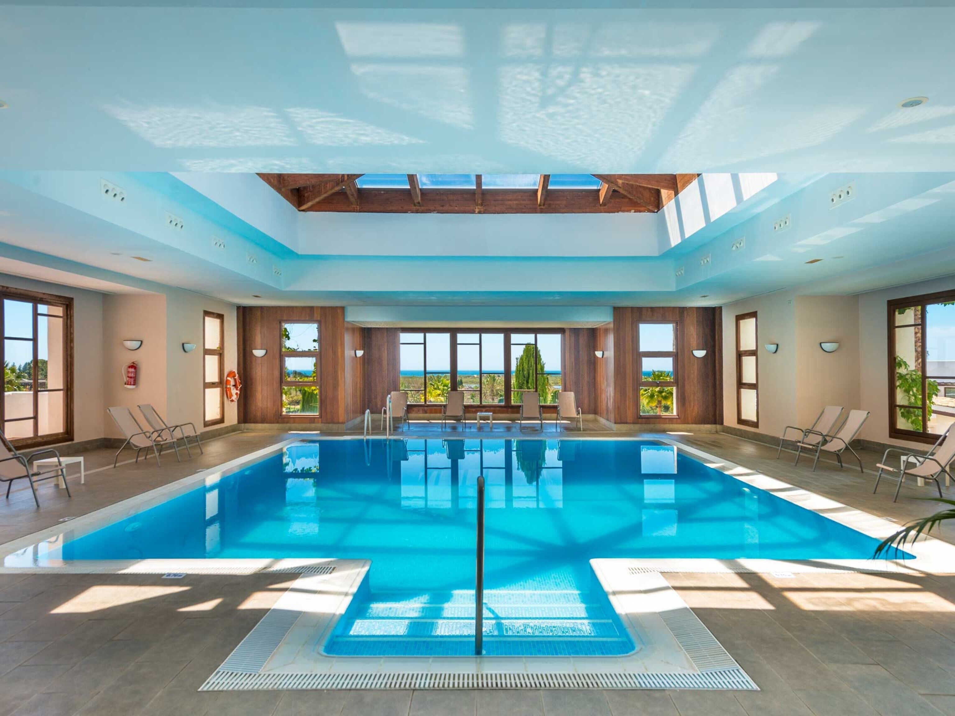 A large indoor swimming pool with clear views of the ocean, wooden ceiling beams, and sunlit windows.