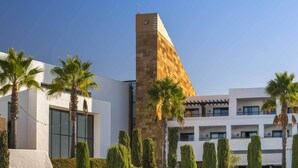 A modern hotel with a prominent tower, palm trees, and neatly trimmed hedges.