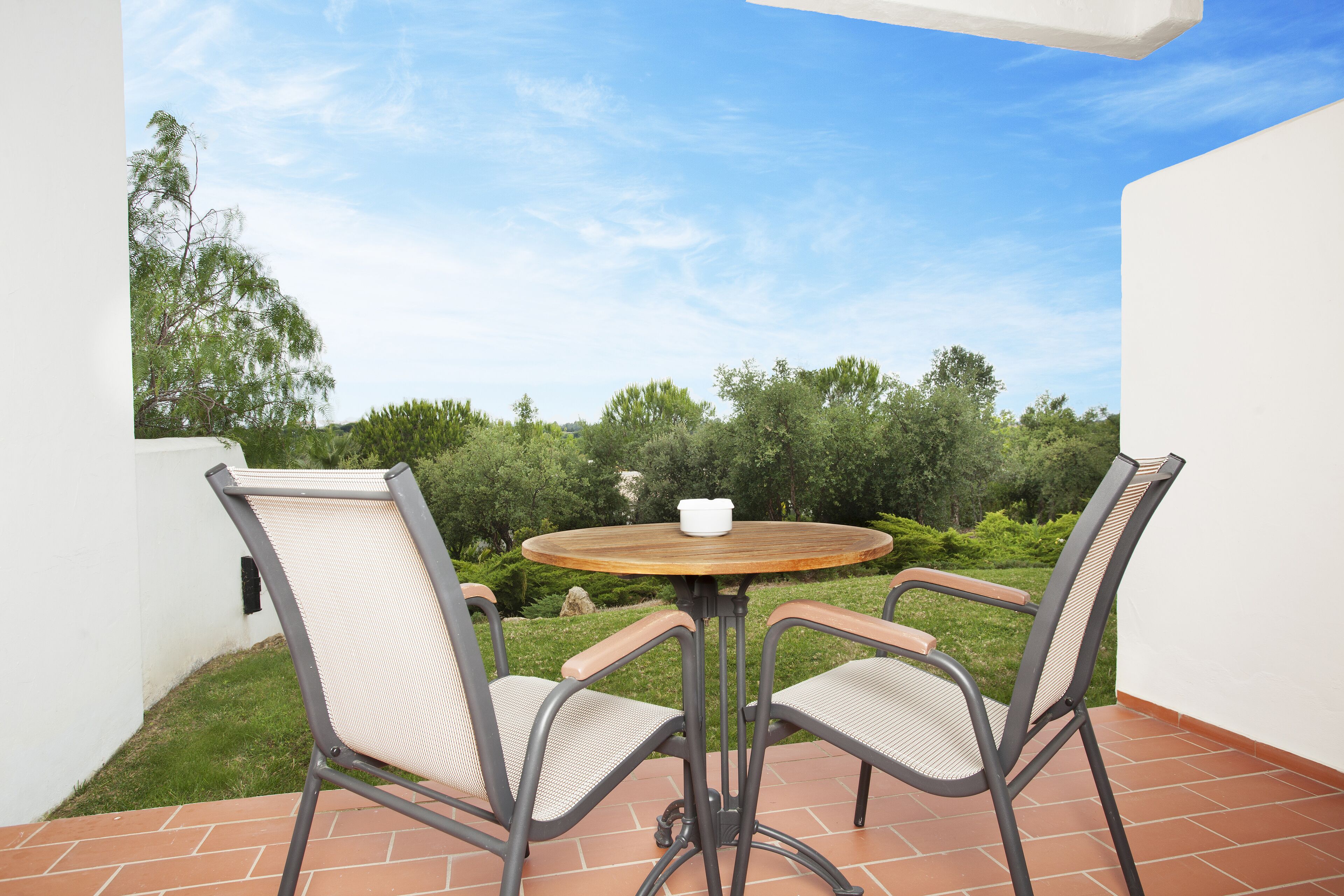 An outdoor patio with a round table and two chairs, surrounded by greenery and a clear sky.