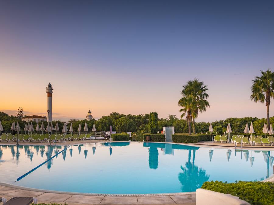 Una piscina con sillas de descanso y sombrillas, rodeada de palmeras y un faro al fondo.