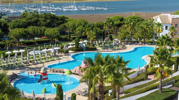 A resort with a large pool, palm trees, and a marina in the background. - Hotel Fuerte El Rompido (Cartaya)