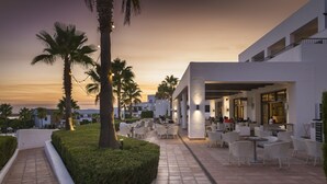 An outdoor seating area with tables and chairs, palm trees, and a building with large windows. - Hotel Fuerte El Rompido (Cartaya)