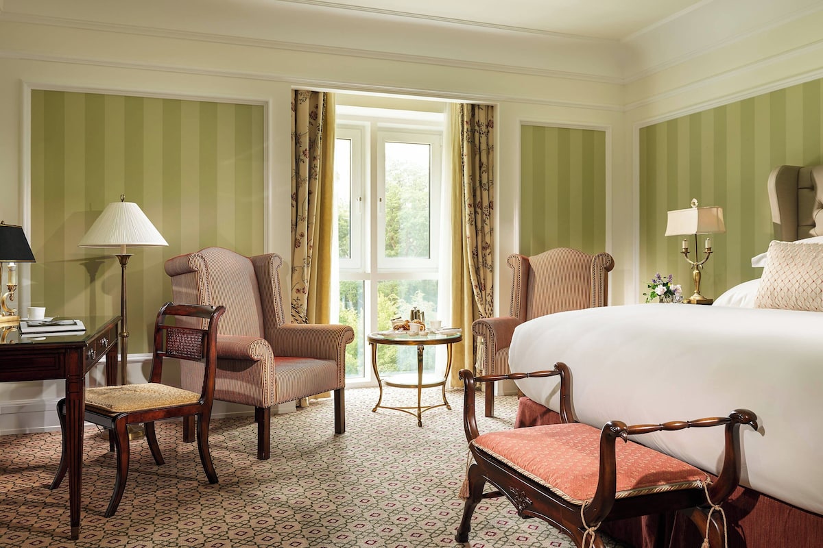 Une chambre d’hôtel avec un grand lit, deux fauteuils, une petite table et un bureau avec une lampe.