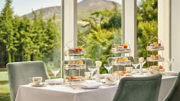 A table set for a tea service with tiered trays of pastries and sandwiches, elegant glassware, and a view of a mountain and greenery.