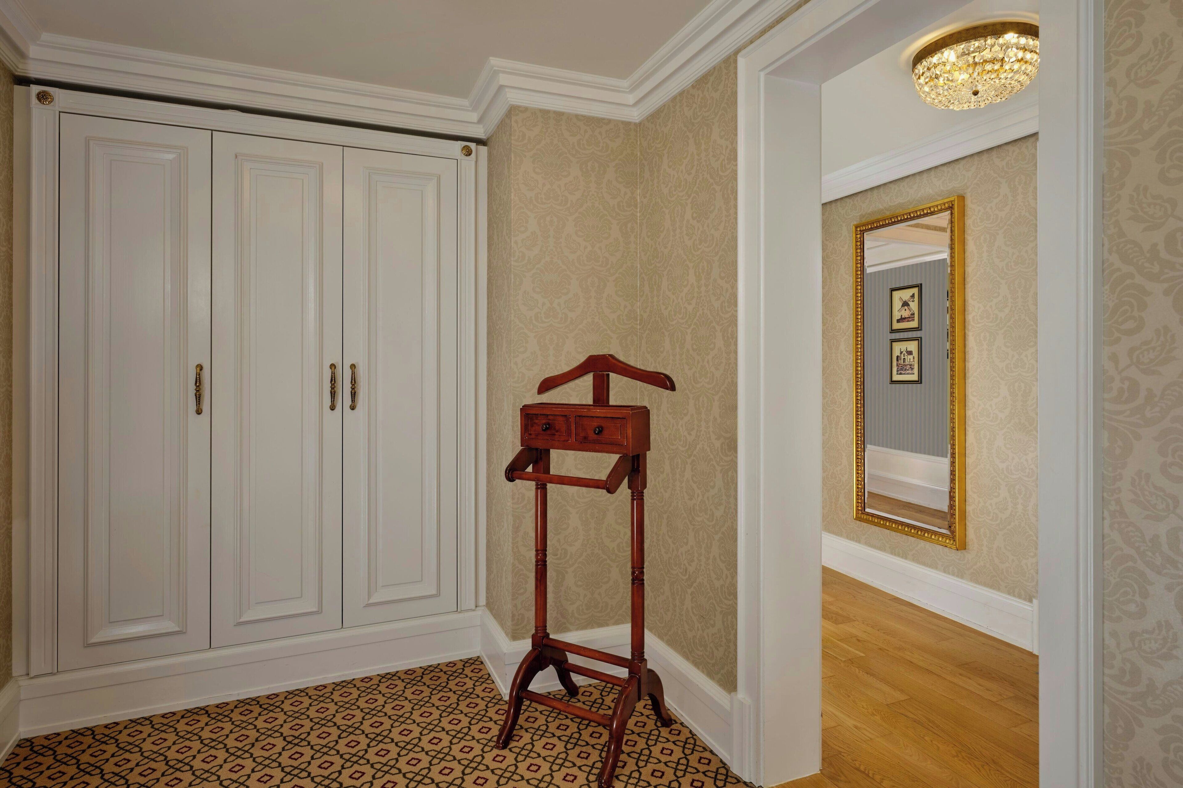 A hallway with a white wardrobe, patterned wallpaper, a wooden stand with drawers, and a mirror with a gold frame.