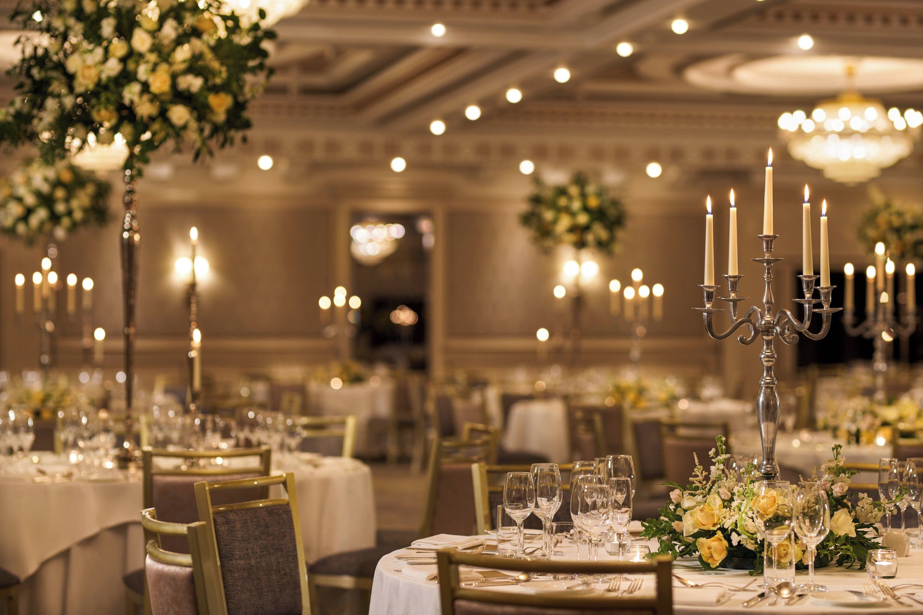 Een feestzaal met ronde tafels gedekt voor een diner, elk met een kandelaar als middelpunt en bloemstukken.