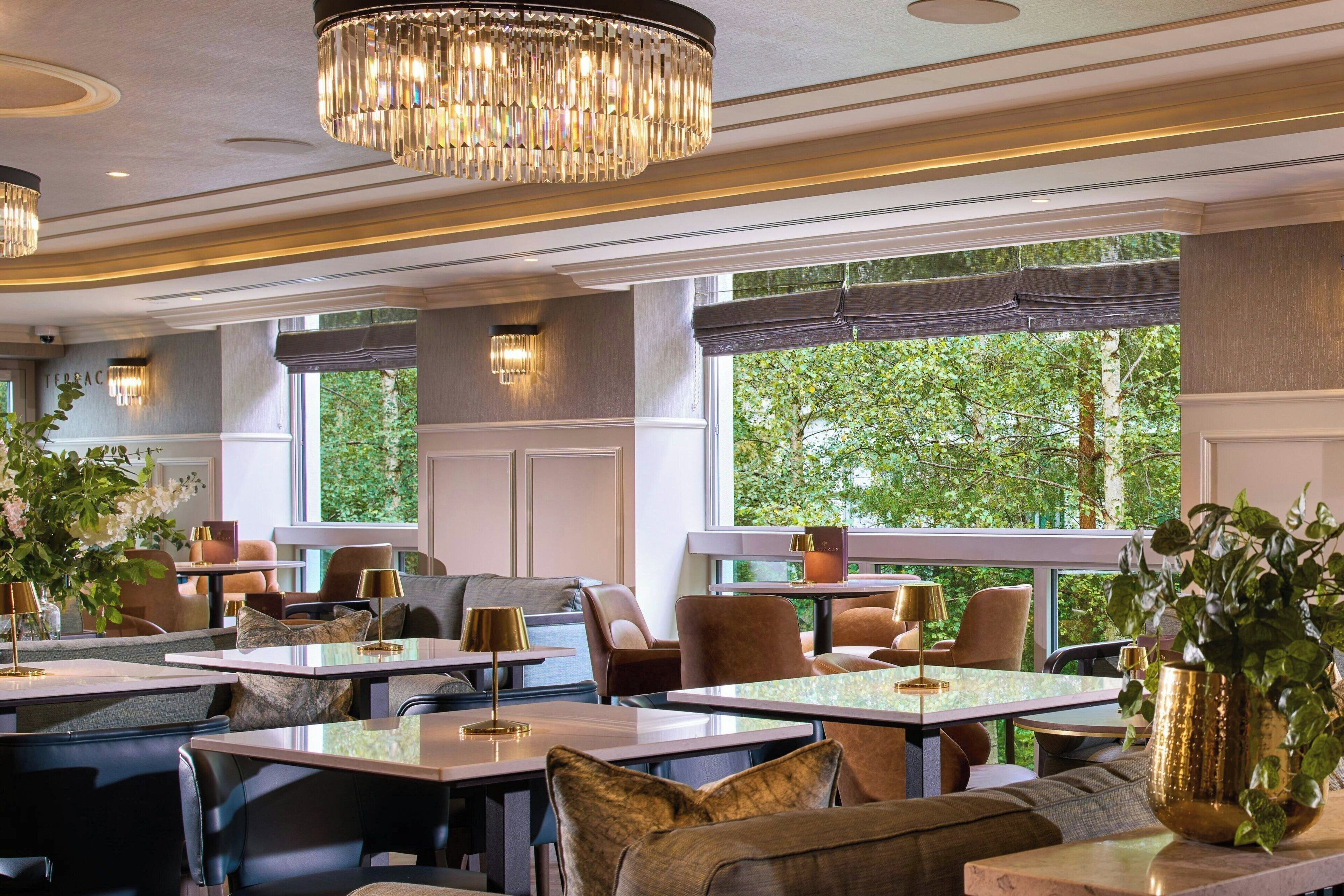 A restaurant with multiple tables and chairs, large windows with a view of greenery, and decorative lighting.