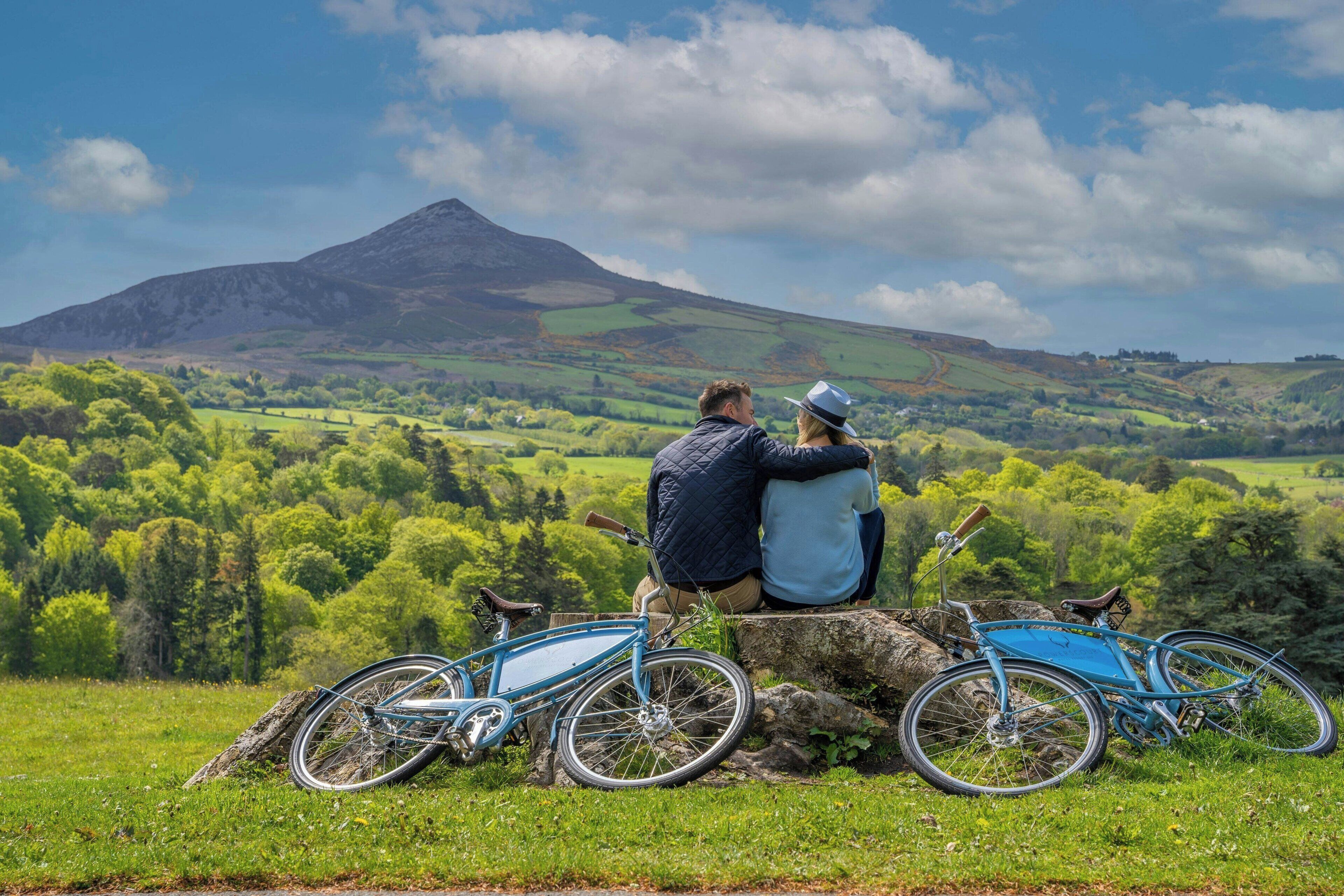 A couple sitting on a rock with two bicycles, overlooking a green valley and a mountain.