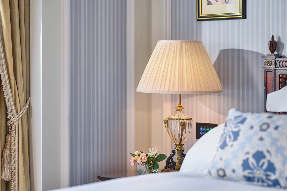A neatly arranged hotel room with a bed, a bedside lamp, a decorative vase with flowers, and a wall-mounted clock.