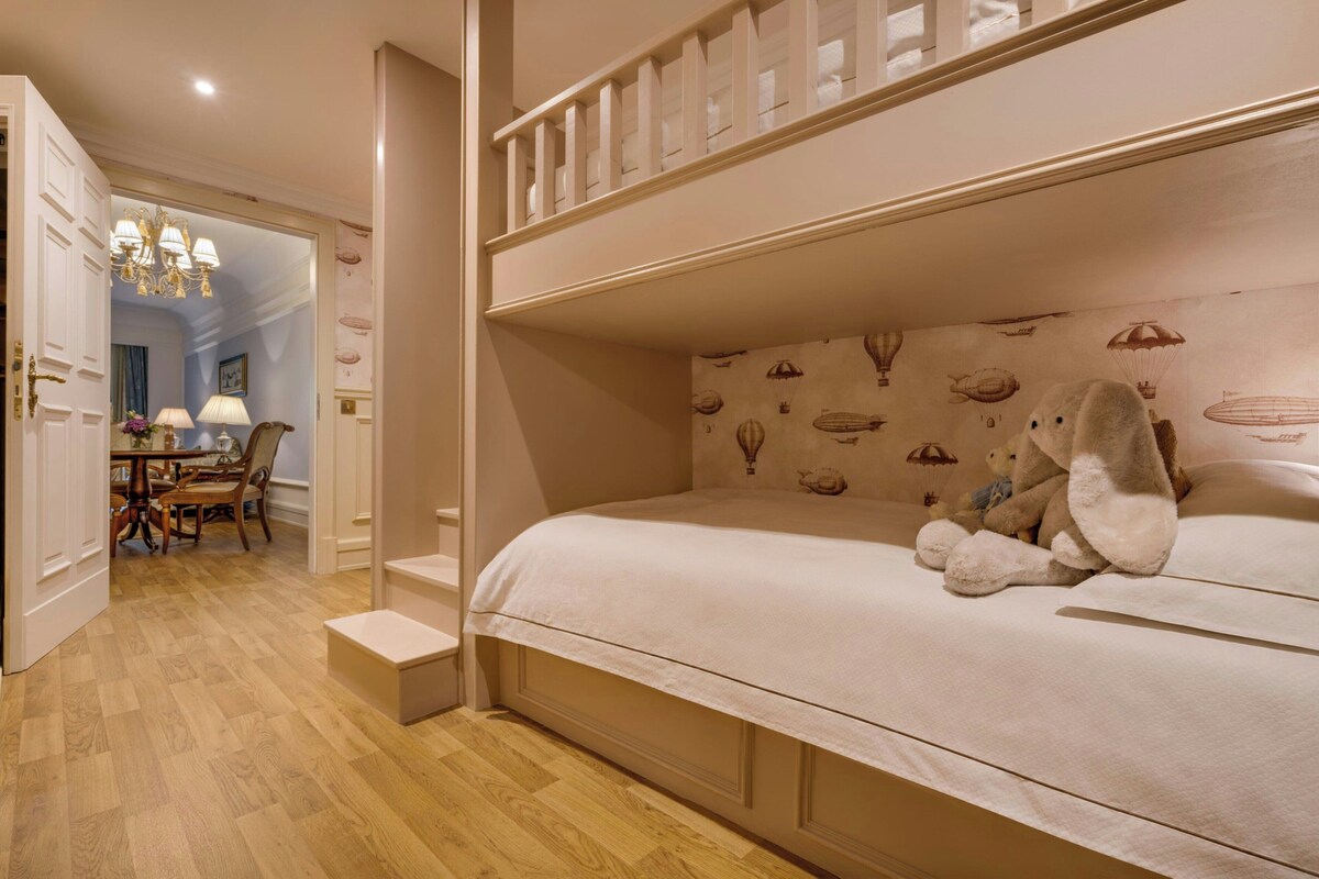 Un dormitorio con una cama litera, un osito de peluche sobre la cama y un papel tapiz con globos aerostáticos.