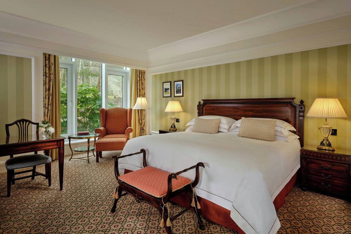 Une chambre d’hôtel avec un grand lit, deux chaises, une petite table et une fenêtre avec des rideaux.