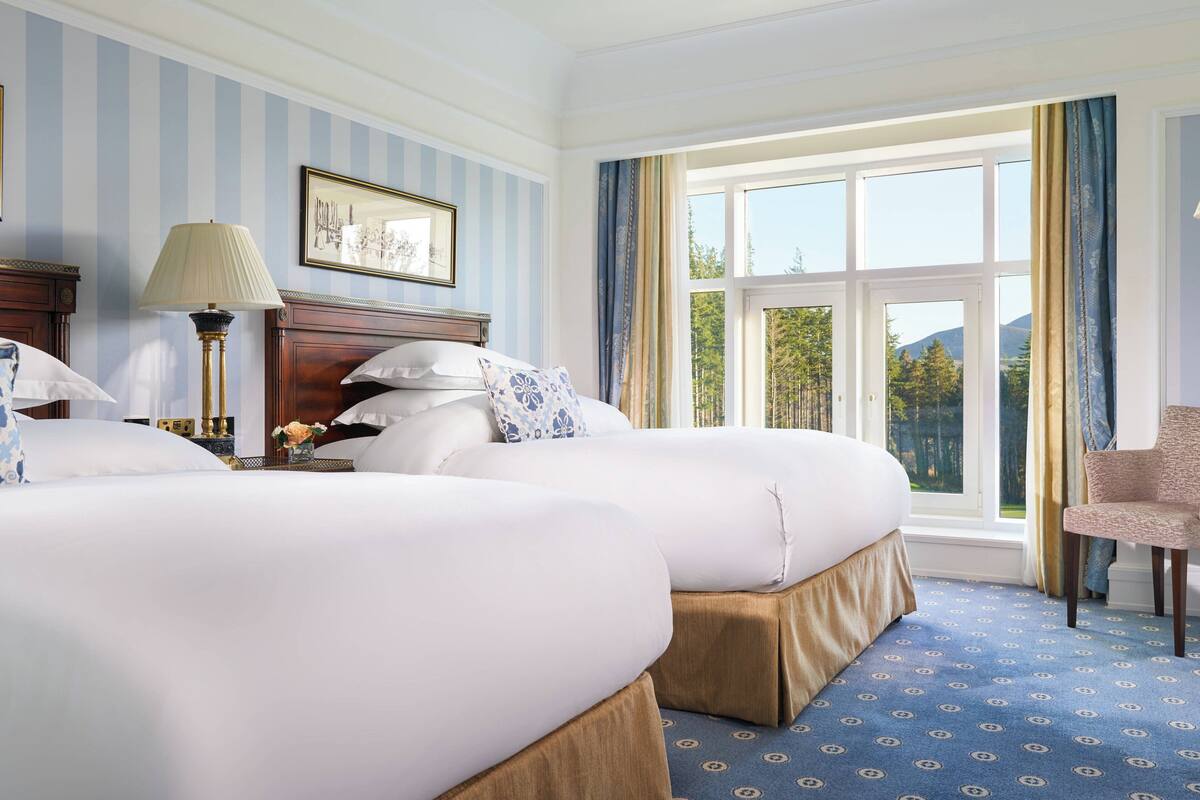 Une chambre d’hôtel avec deux lits, une grande fenêtre avec des rideaux, une lampe et une chaise.