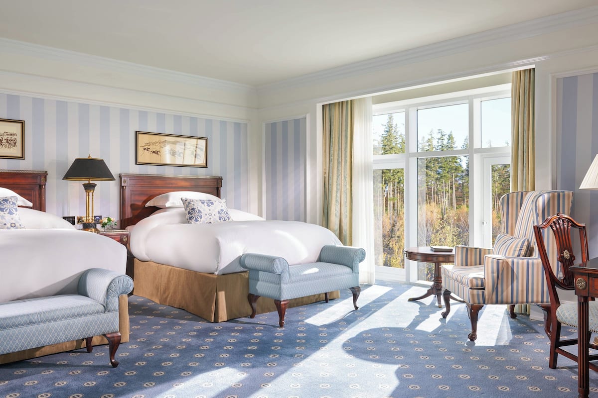 Une chambre d’hôtel avec deux lits, un coin salon comprenant une chaise et une table, et de grandes fenêtres donnant sur la verdure.