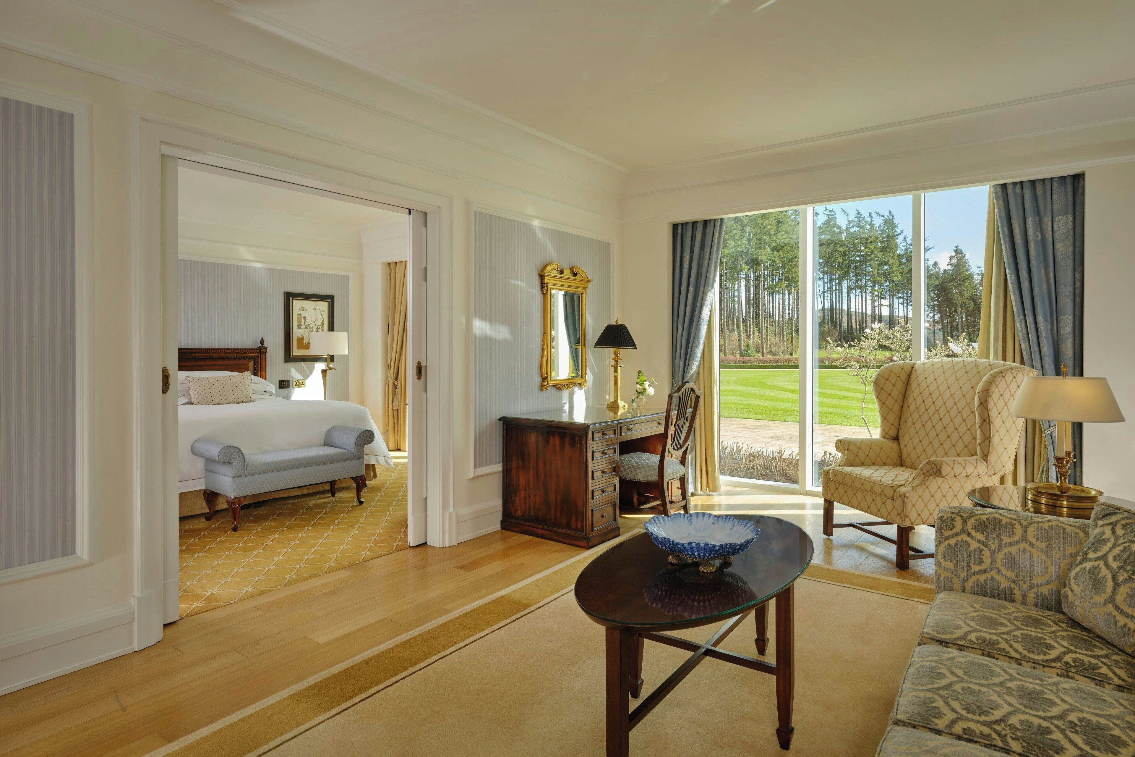 A hotel room with a bed, a sitting area with a sofa and armchair, a dining table, and a view of a golf course through large windows.