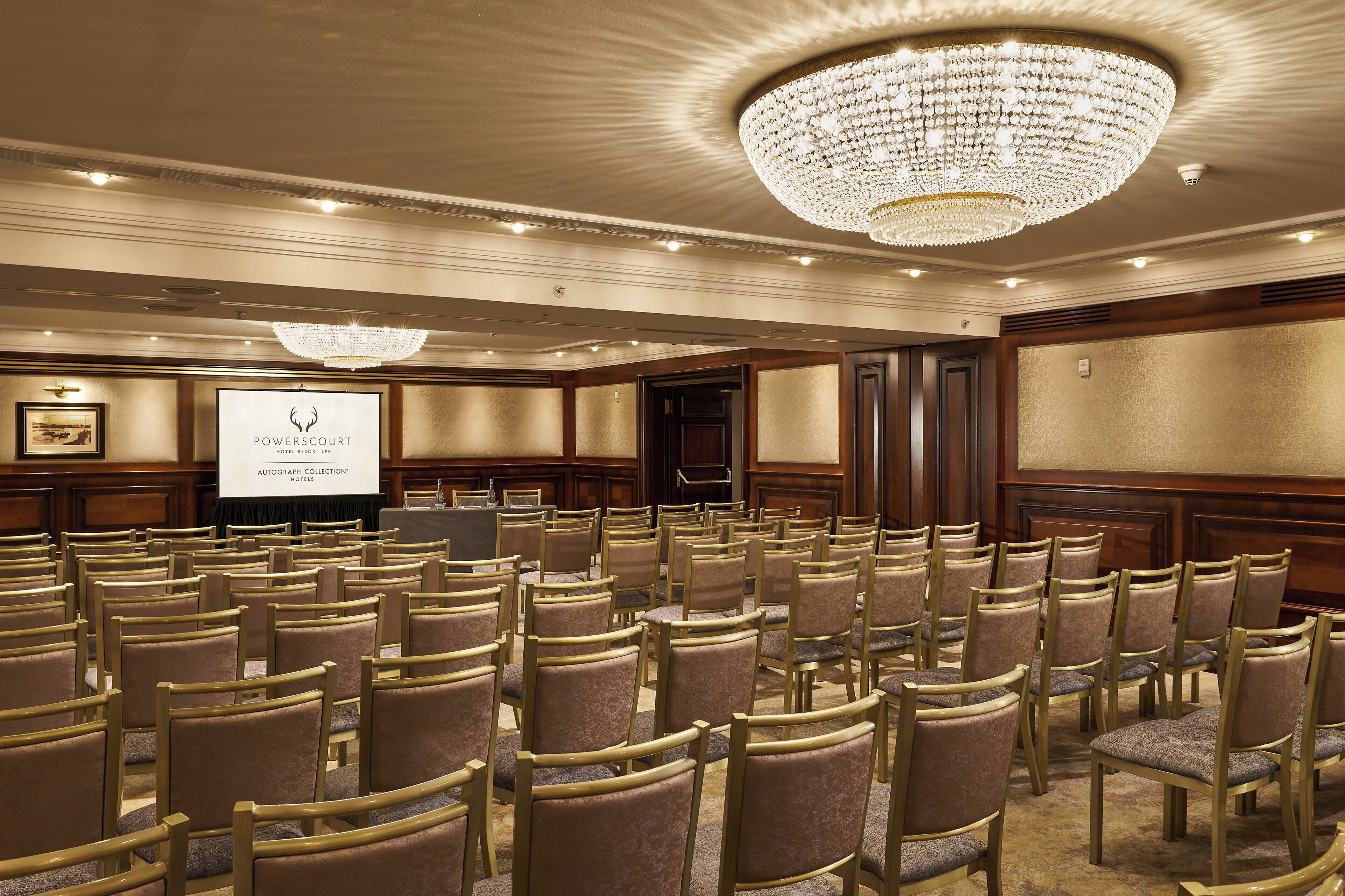 A conference room with rows of chairs, a projector screen, and a chandelier.