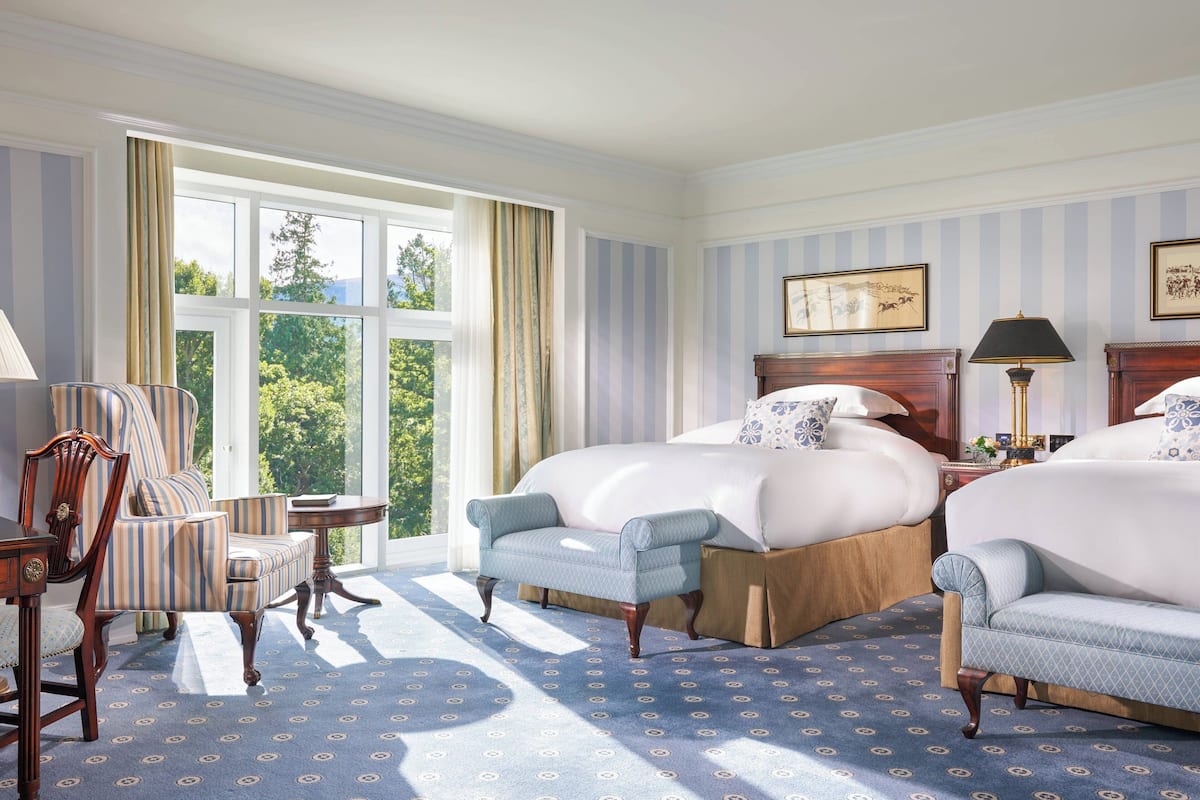 Une chambre d’hôtel avec un grand lit, deux fauteuils, une chaise en bois, une petite table et une fenêtre avec des rideaux.